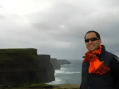 Rob Tatum smiling near dramatic seaside cliffs under a cloudy sky, wearing sunglasses and a bright orange scarf.