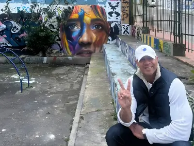 Rob Tatum crouching and smiling with a peace sign beside a colorful mural in an urban outdoor space.