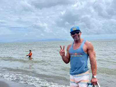 Rob Tatum standing at the beach, smiling and flashing a peace sign under a cloudy sky.