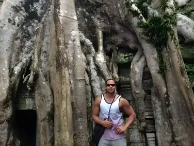 Rob Tatum standing in front of massive tree roots and ancient stone ruins, wearing sunglasses and a tank top.