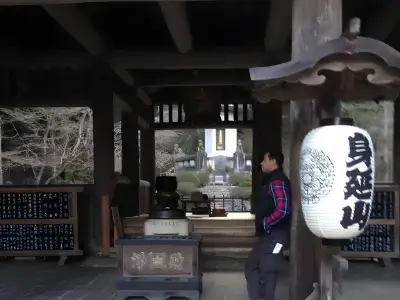 Person standing inside a traditional Japanese temple entrance beside a large hanging lantern, with a courtyard visible in the background.