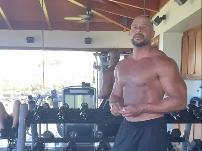 Rob Tatum standing shirtless in a home gym, with dumbbells and workout equipment in the background.