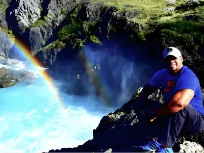 Rob Tatum sitting on rocky cliffs beside a turquoise waterfall with a rainbow in the mist.