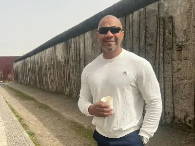 Rob Tatum smiling and holding a cup while standing beside a historic concrete wall outdoors.