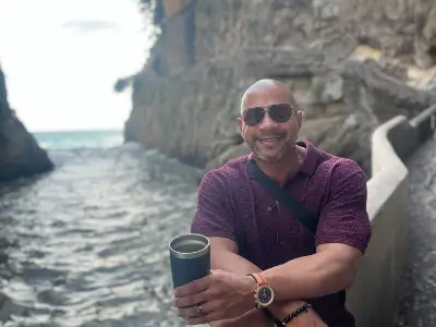 Rob Tatum smiling and holding a tumbler while standing beside a rocky coastal walkway.