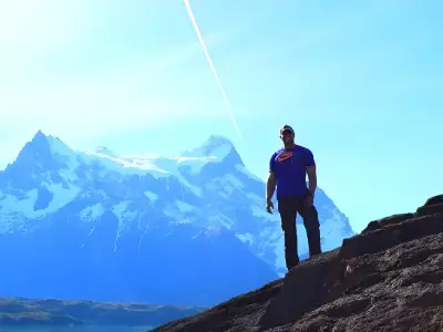 Rob Tatum standing on a rocky slope with snow-capped mountains and a bright blue sky in the background.