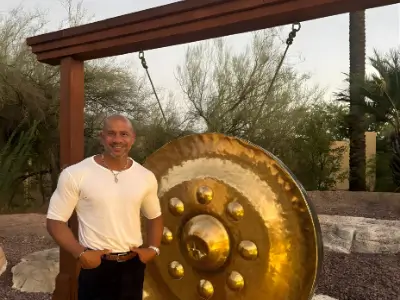 Rob Tatum standing beside a large golden gong outdoors under a wooden frame, smiling at the camera.