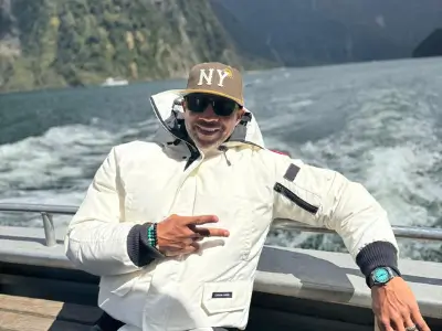 Rob Tatum posing on a boat in a white jacket and NY cap, flashing a peace sign with mountains and water in the background.