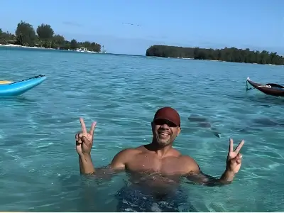 Rob Tatum smiling in clear turquoise water, holding up peace signs with small islands and boats in the background.