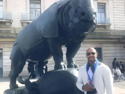 Rob Tatum smiling beside a large bull statue in a city plaza, wearing sunglasses and a white jacket.
