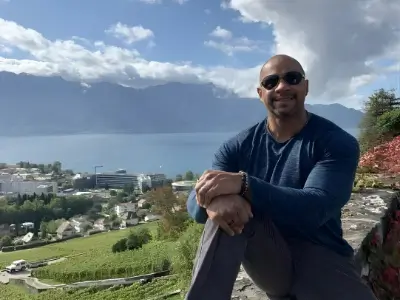 Rob Tatum sitting outdoors on a hillside overlook with mountains and a lake in the background.