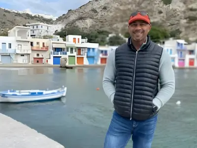 Rob Tatum smiling by a waterfront harbor, wearing a black puffer vest and red cap, with colorful boathouses and rocky hills in the background.