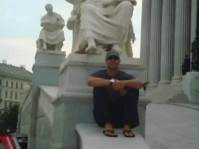 Rob Tatum sitting on marble steps near large classical statues and columns, wearing sunglasses, a dark shirt, and sandals.