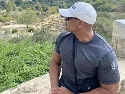 Rob Tatum sitting outdoors on a stone ledge, wearing a white cap and sunglasses, overlooking a green hillside landscape.