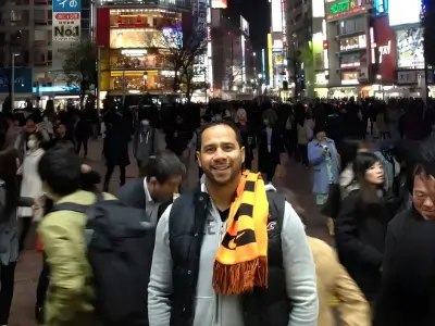 Rob Tatum smiling in a busy city street at night, wearing a black-and-orange scarf amid a crowded urban scene with bright neon signs.