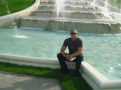Rob Tatum sitting by a large cascading fountain on a sunny day, wearing sunglasses and casual dark clothing.