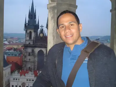 Rob Tatum smiling beside a historic city skyline, with gothic towers and red rooftops visible in the background.