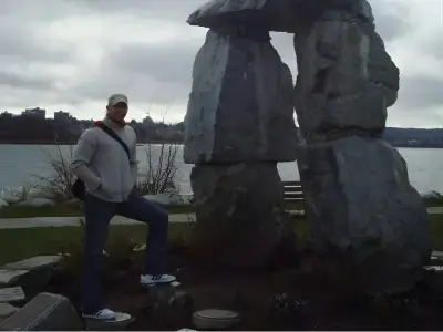 Rob Tatum standing beside a large stone sculpture near a lakeside under a cloudy sky.