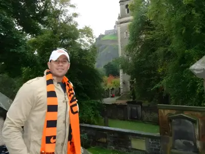 Rob Tatum smiling outdoors in a white cap and orange striped scarf near a historic stone structure surrounded by trees.