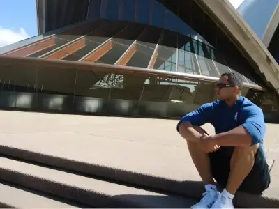 Rob Tatum sitting on outdoor steps in front of a modern glass building, wearing sunglasses and a blue shirt on a sunny day.