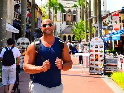 Rob Tatum smiling and walking through a sunny outdoor shopping district with palm trees, wearing a navy tank top and backpack.
