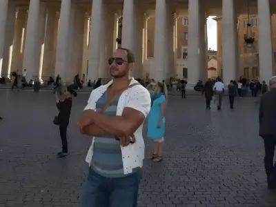 Rob Tatum standing with arms crossed in a large public square with towering columns, wearing sunglasses and casual summer clothes.