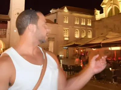 Rob Tatum gesturing while standing in a warmly lit outdoor plaza at night, wearing a white tank top and crossbody bag.