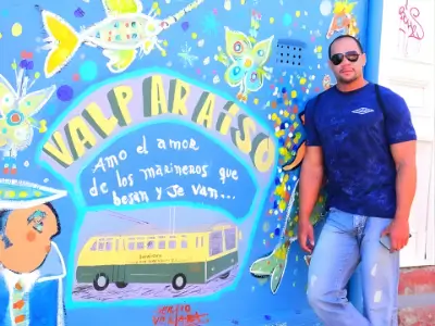 Rob Tatum standing beside a colorful mural with playful artwork and Spanish text, wearing sunglasses, a blue shirt, and jeans.