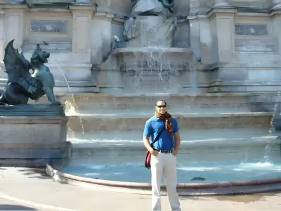 Rob Tatum standing in front of a large fountain with cascading water and sculptural details, wearing sunglasses, a blue shirt, and light-colored pants.