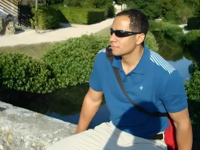 Rob Tatum wearing sunglasses and a blue polo shirt, leaning on a stone wall beside a stream on a sunny day.