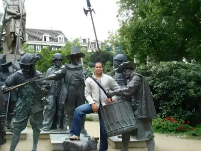 Rob Tatum posing among bronze soldier statues in a park memorial, smiling with one foot raised beside a drum sculpture.